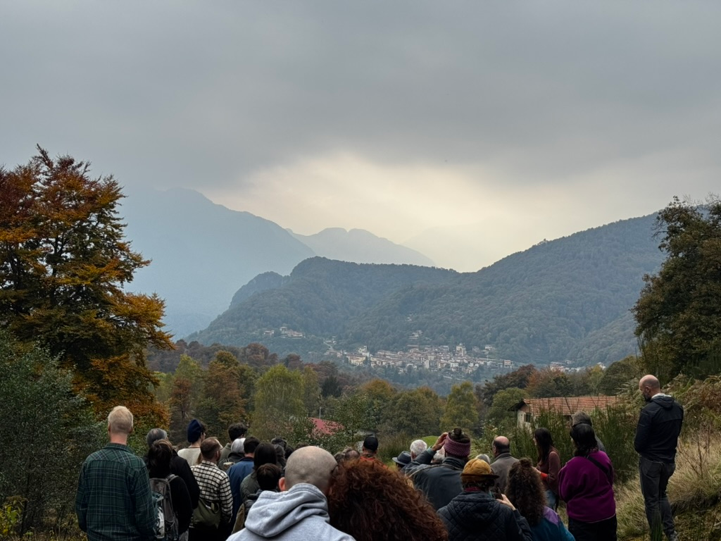 Crowd of people gathered outdoors facing mountain village nestled in valley, with layered mountain ranges and overcast sky in background.
