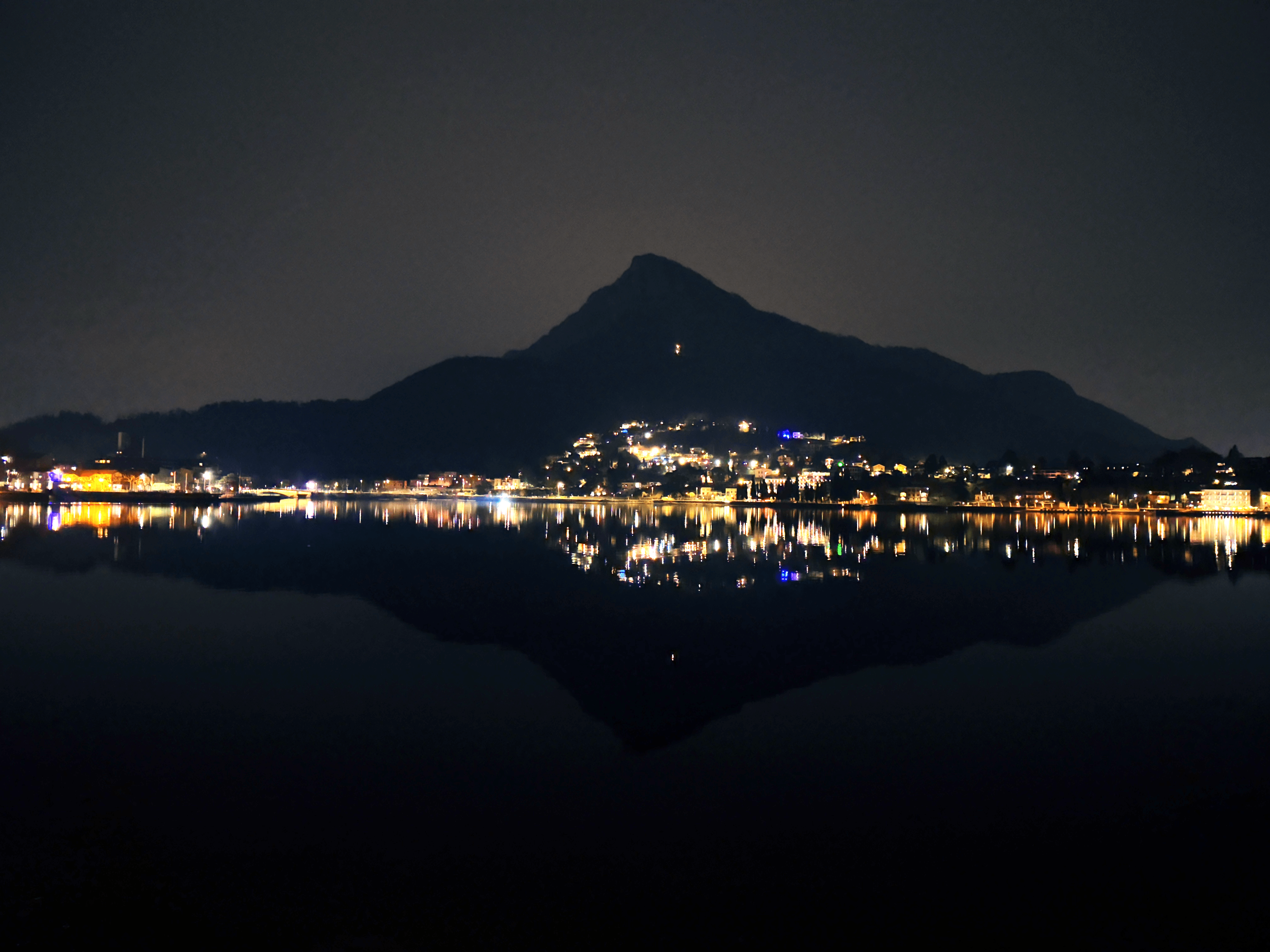 The lights of a city at night and the shadow of the mountain behind it are perfectly reflected in the dark waters of the lake.